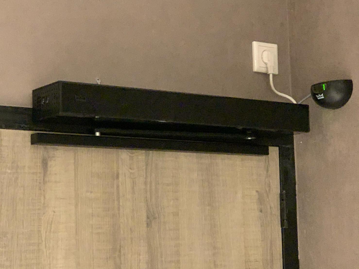 Zwarte deurautomaat boven een houten deur, met een sensor naast de deur